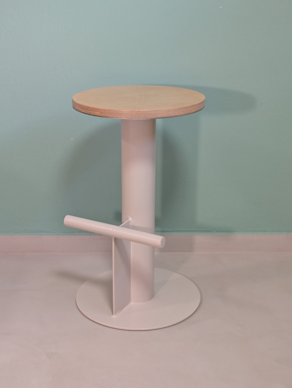 A single finished barstool with a plywood seat and white steel base, against a pale green wall
