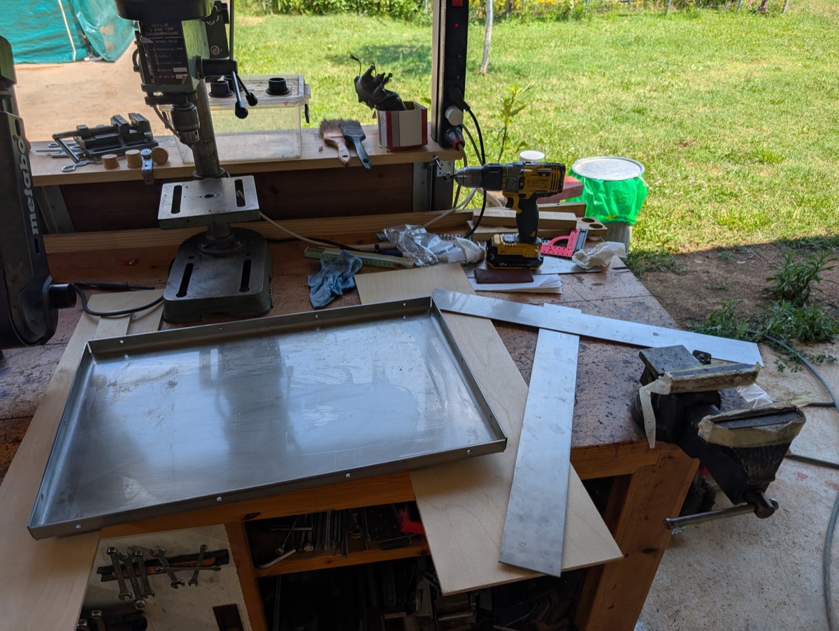 A drilled steel tray on the workbench beside a drill press and hand drill